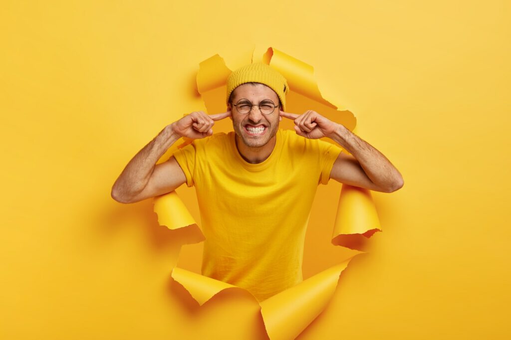 Irritated man plugs ears with both hands, ignores bad sound, wears yellow hat and t shirt, stands in